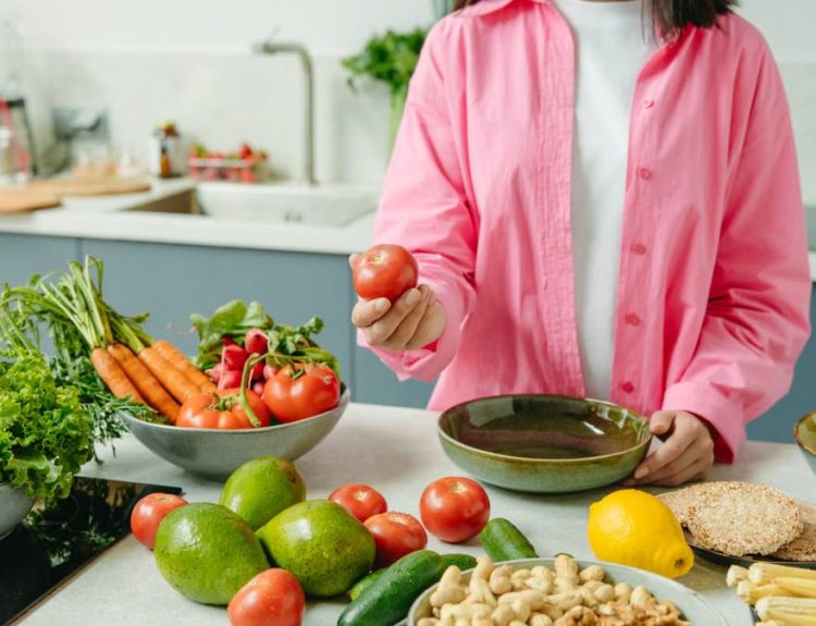 Woman in pink overshirt holds tomato in kitchen with fresh fruits and veggies.