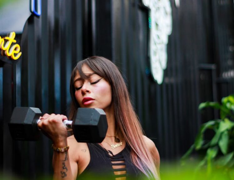 Fit woman with long hair and tattoos lifting a dumbbell with intense focus outdoors.